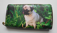 Pug Wallet 11. Grass and plant green background, cute Pug with exposed tongue and aux leather zip-around with zippered coin sleeve as a divider and five card slots. Wallet Measures 7.50" X 4.50" (19.05 X 11.43 cm)